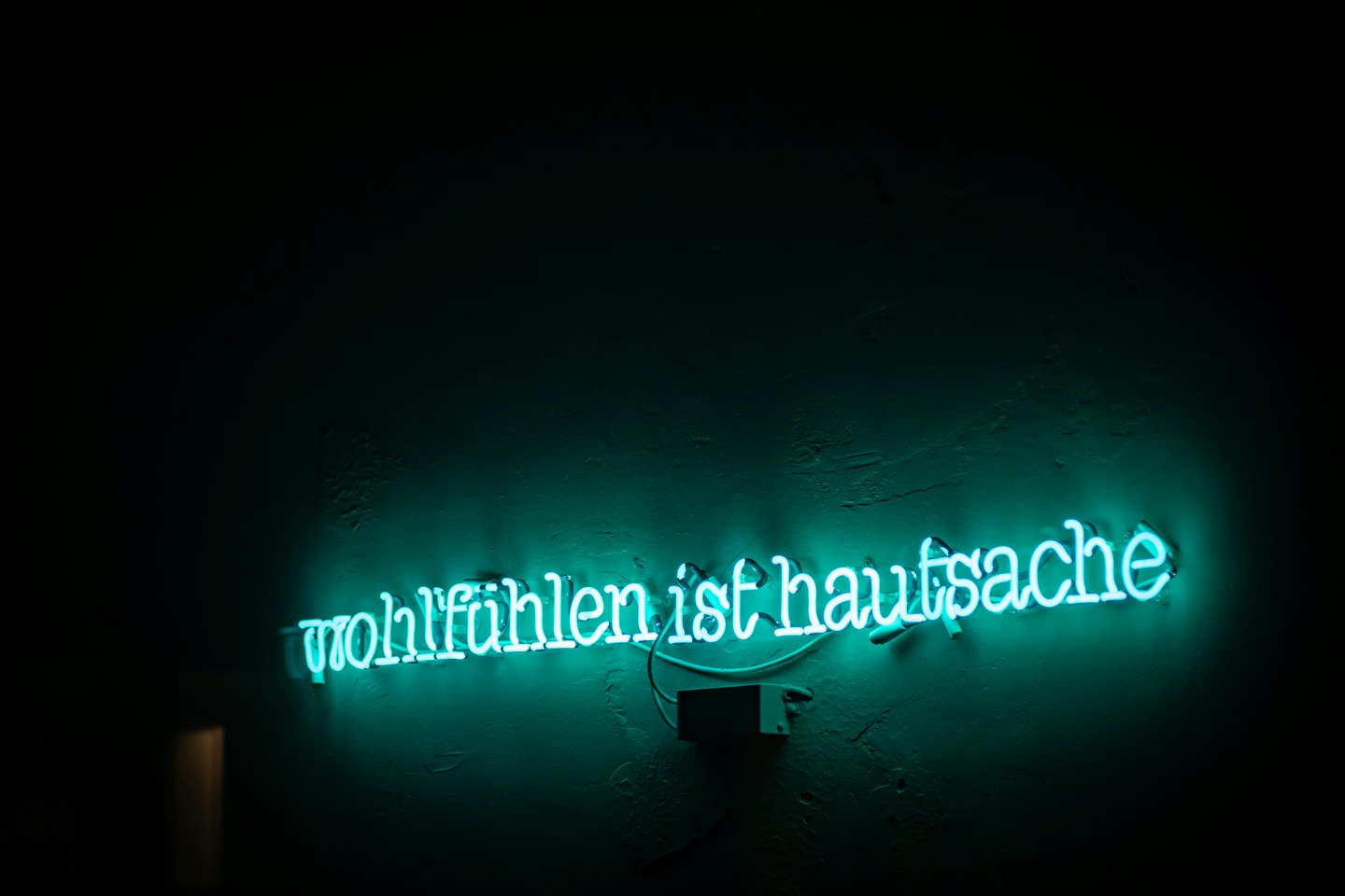 Türkises Neonschild mit der Aufschrift 'Wohlfühlen ist Hauptsache' an einer dunklen Wand.